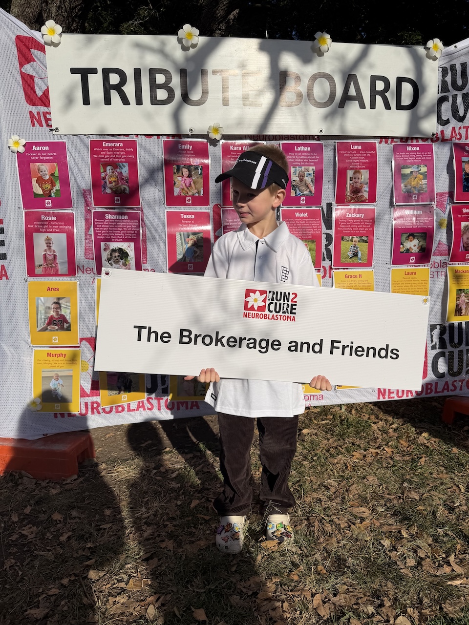 A young supporter holding The Brokerage and Friends team sign in front of the Run2Cure tribute board